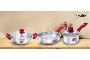Prestige stainless-steel cookware set, including a frying pan and a saucepan with red handles, displayed with kitchen utensils.